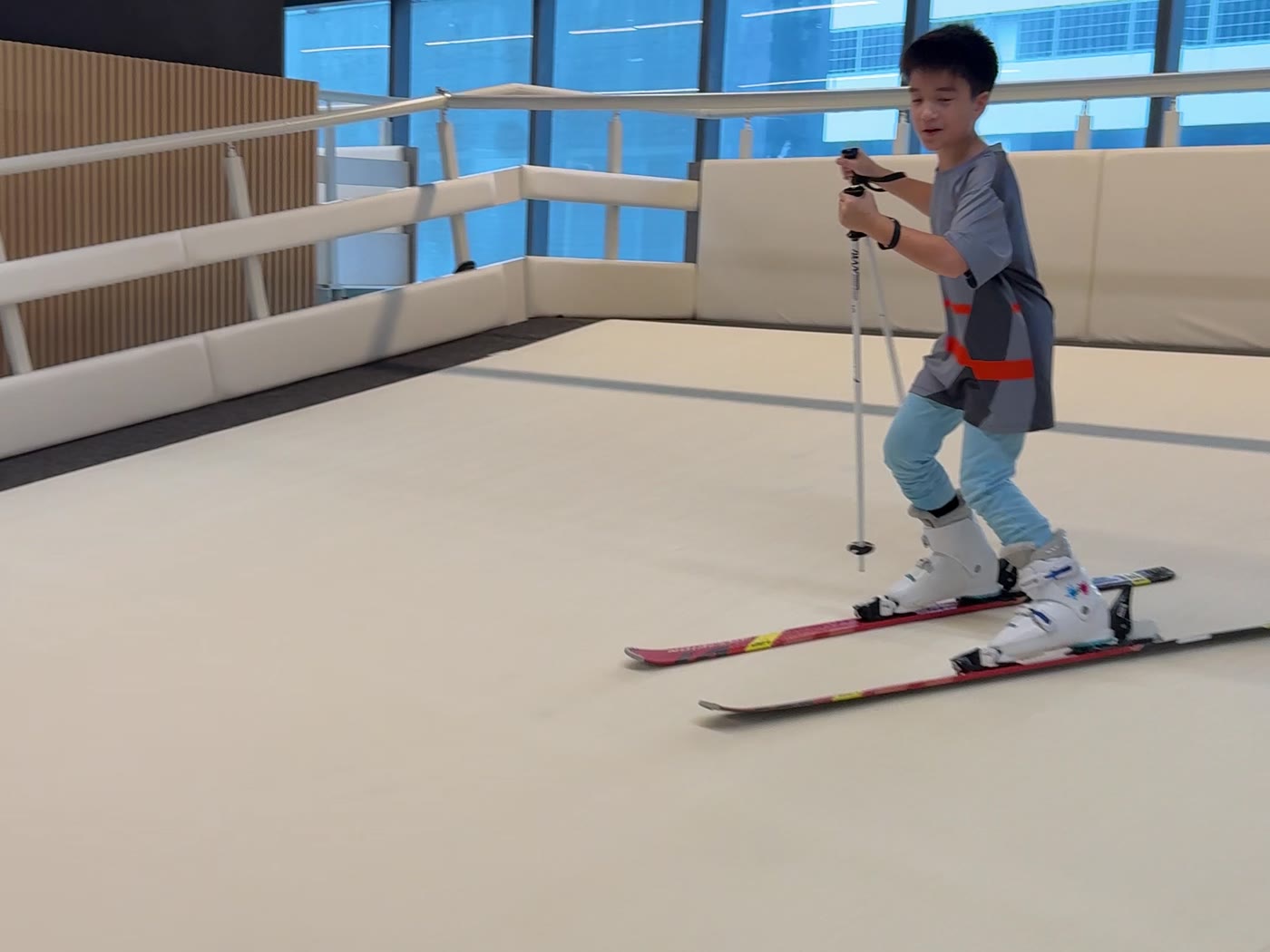 A child skiing with poles on the moving slope at SKI Academy Thailand, Bangkok skyline through the glass
